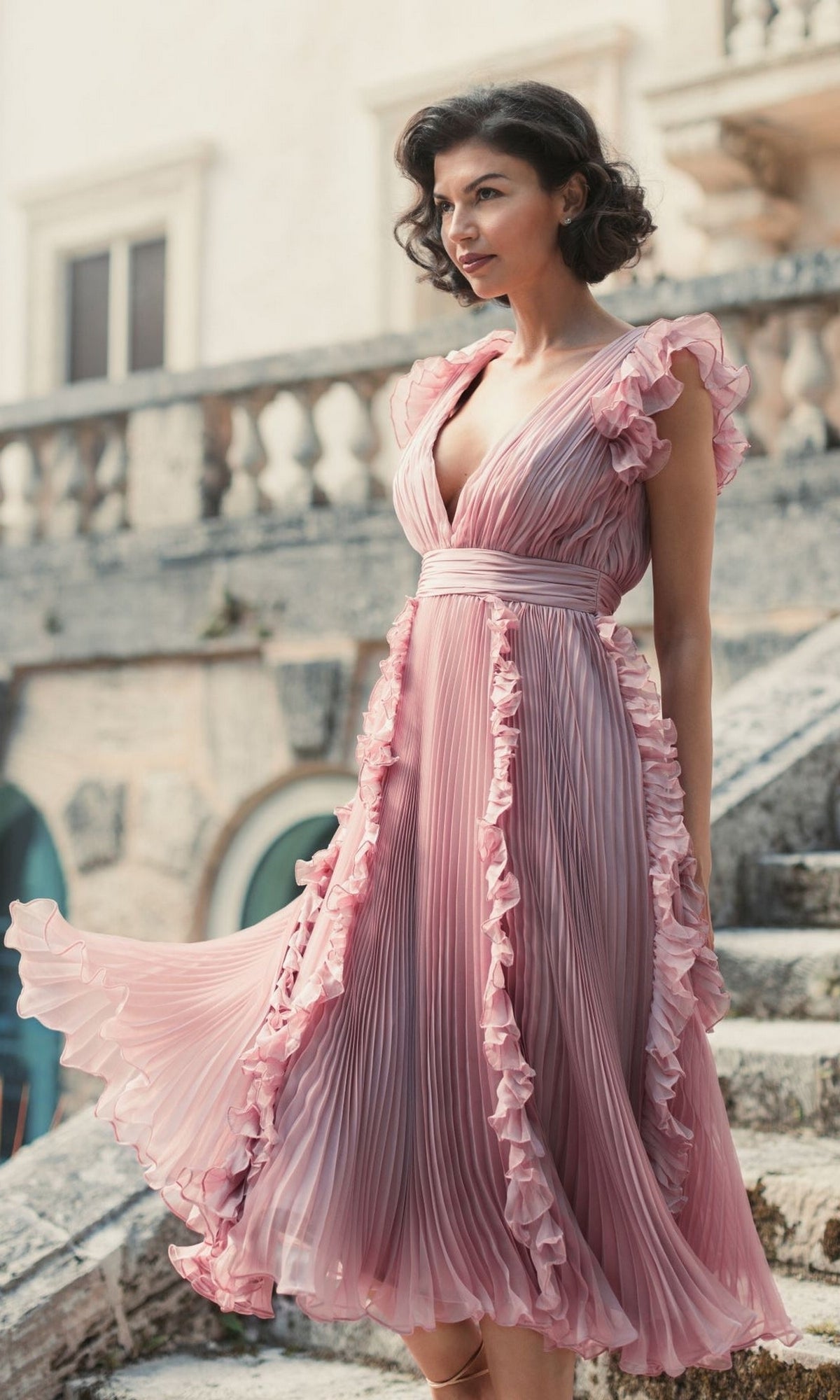 A woman in the rose pink Short Wedding Guest Dress 49675 by Mac Duggal, featuring ruffle cap sleeves, stands outdoors on stone steps with a balustrade and building behind her.